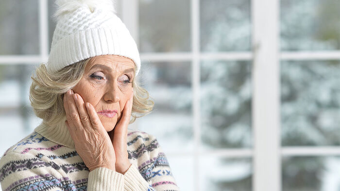 Image of an elderly woman wearing a warm sweater and wool hat indoors. She looks worried and sad.