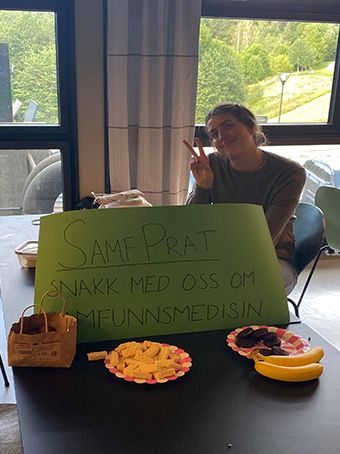 Anne Sigrid med en grønn plakat med "Snakk med oss om samfunnsmedisin", På borde ligger diverse snacks.