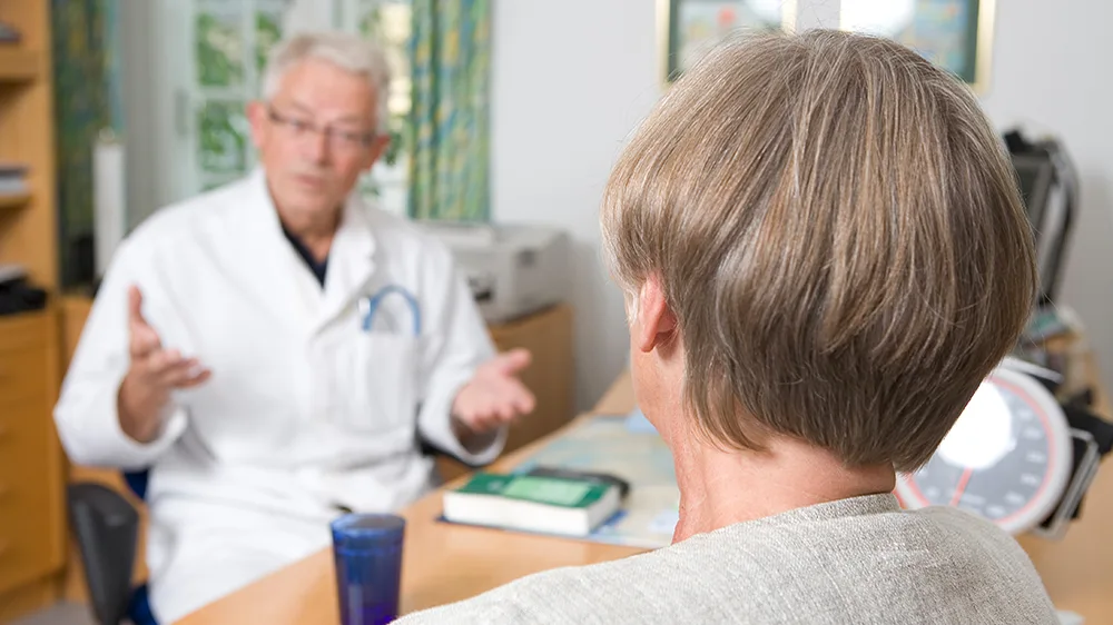 En lege som sitter ved et skrivebord og snakker til en pasient.