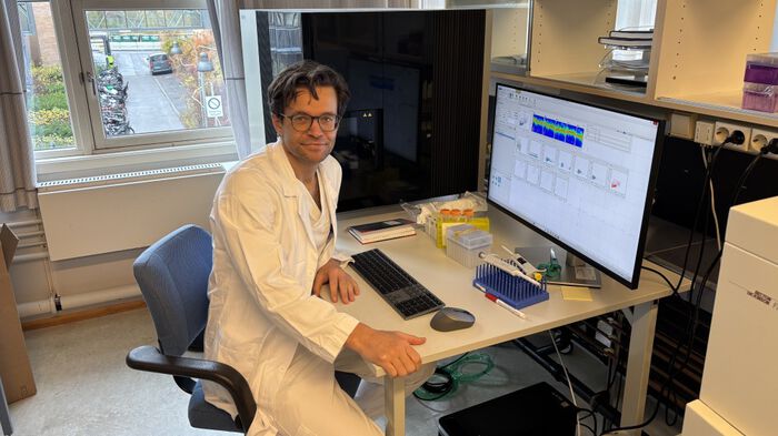 Image shows a man witg glasses in front of a black machine connected to a computer
