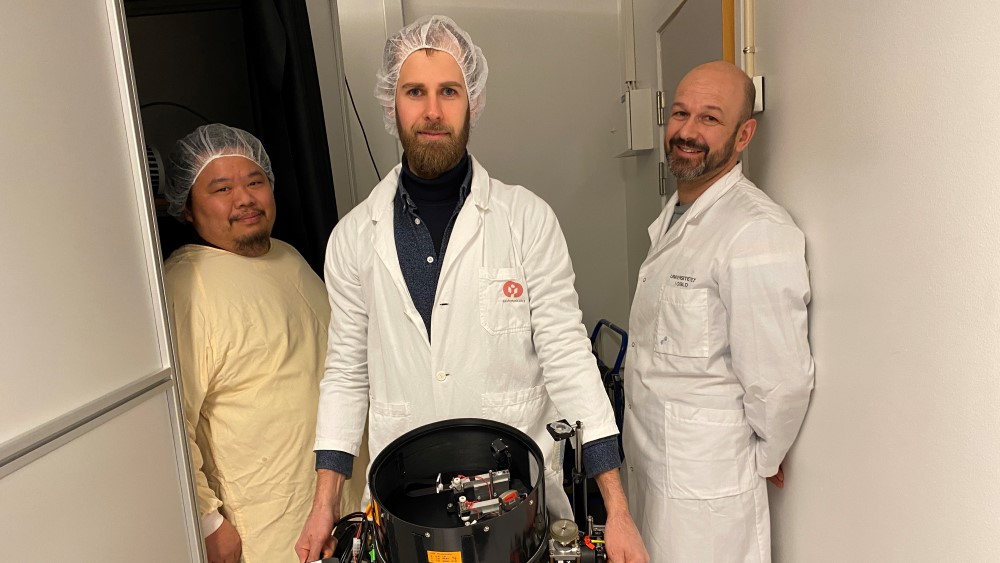 Bildet viser tre menn i labfrakker og med hette på hodet, som holder i et svart, rundt intrument med ledninger og sylindere
