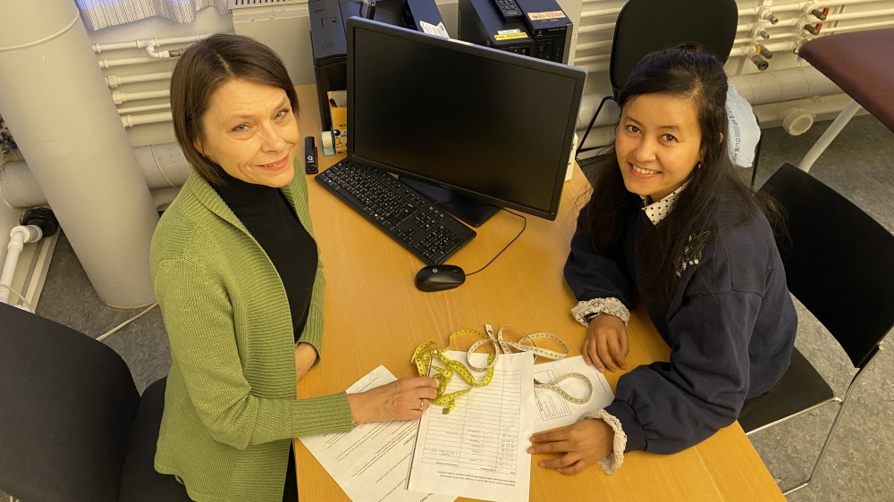 Bildet viser to kvinner som sitter ved et skrivebord med skjemaer og et målebånd.