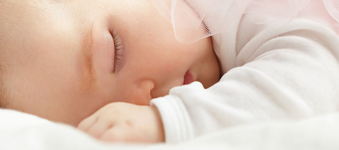 Nærbilde av et sovende barn, med en arm foran ansiktet. Iført hvite klær.