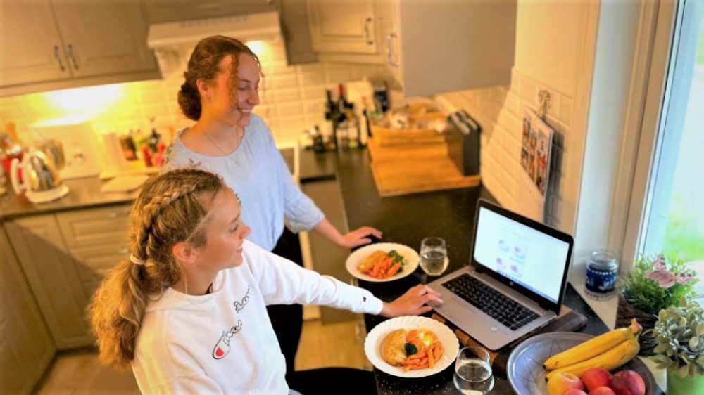 Bildet viser to jenter som sitter med mat foran en PC på et kjøkken