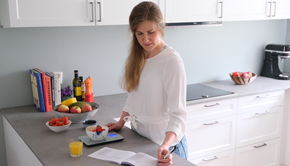 Bildet viser en kvinne på et kjøkken som veier mat og fører noe opp i en bok,