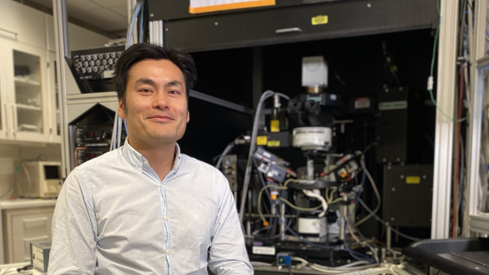 Evandro Fang sitting in front of expensive brain research equipment. He is wearing a blue shirt and is smiling.