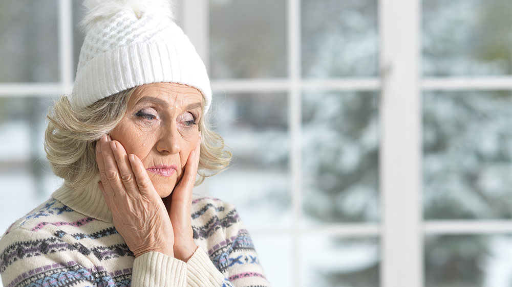 Bilde av en eldre kvinne som har på seg lue og varm genser innendørs og ser bekymret ut