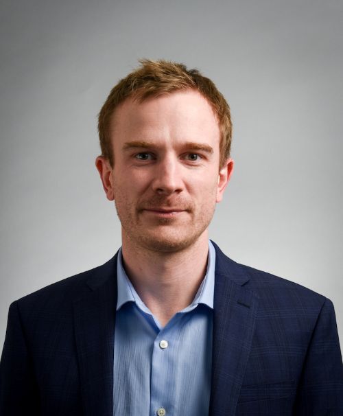 Headshot of David Knowles on a gray background, he is wearing a blue shirt with a blue suit jacket.
