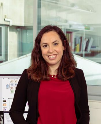 Photo of Laura Cantini wearing a red jumper under a black jacket. She is standing in front of a computer screen and a window.