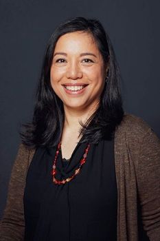 Portrait of Camila Esguerra. She is wearing a black shirt, a brown cardigan and a red beaded necklace. She has shoulder length dark hair and is smiling. 