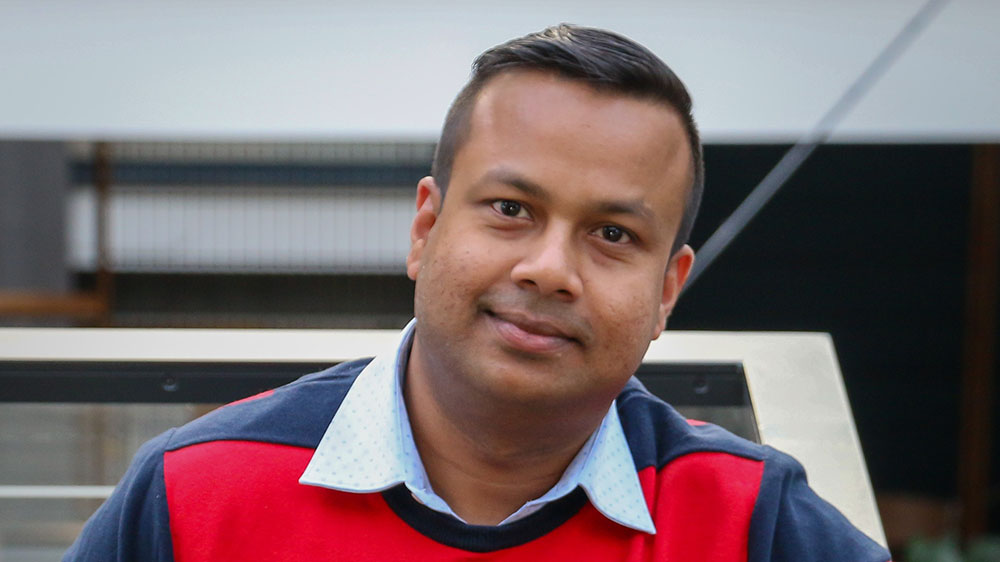 Portrait of Biswajyoti Sahu. He is wearing a red and navy striped shirt and is posing in front of a building