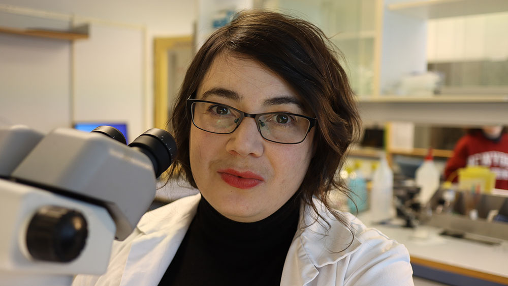 Photo of Charlotte Boccara sitting in a lab. She is wearing a white lab coat, a black polo neck and has shoulder length black hair, wears glasses and has red lipstick.