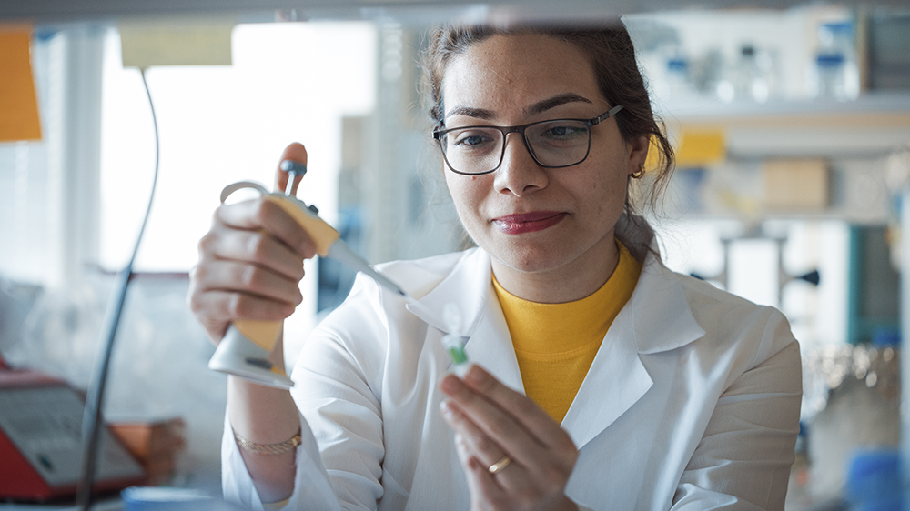 Elham Shojaeinia in a lab. She is pipetting into a tube. She wears a yellow top, a white lab coat and glasses. She has dark hair.