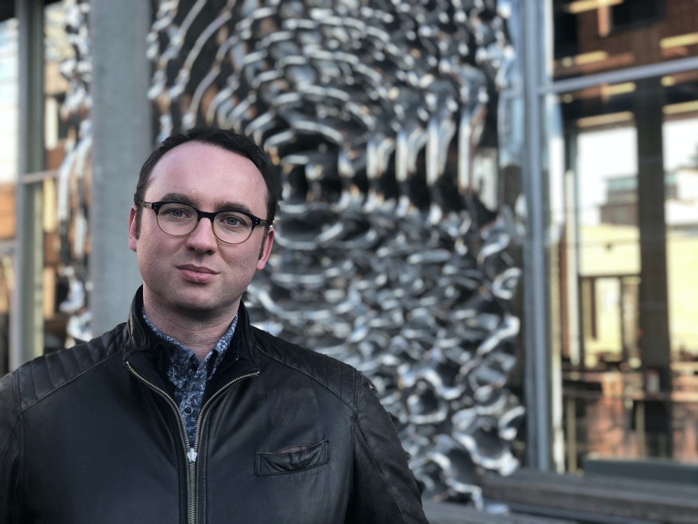Sebastian Waszak standing outside of a building with a metallic sculpture behind him. He's wearing a dark motorcycle jacket, a blue shirt and dark, round glasses