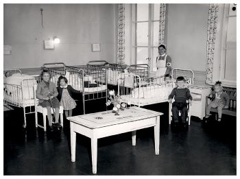 Svart.hvitt-foto fra en sykesal. Fire barn sitter på hver sin stol i forkant av sengene. En sykepleier står bak barna.