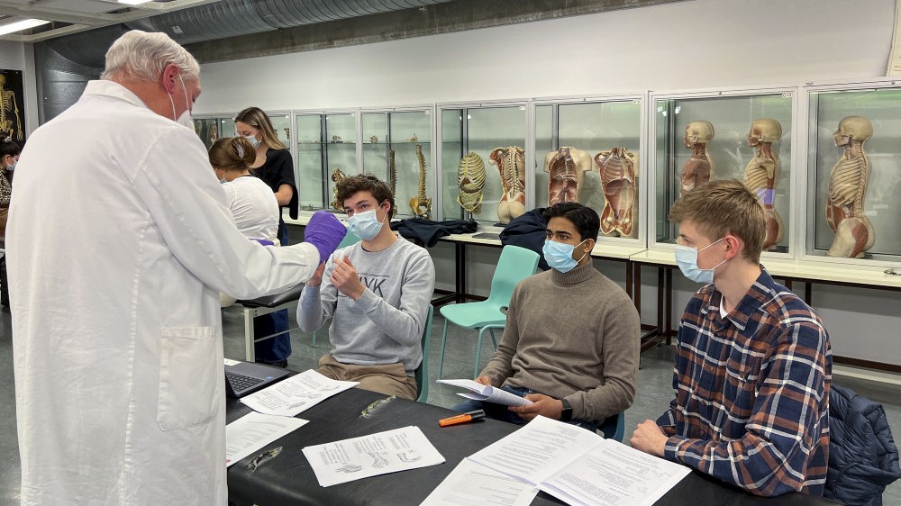 Bildet viser en mann i hvit frakk, plasthansker og munnbind som viser noe til tre studenter med munnbind som sitter ved pulter med en rekke papirer