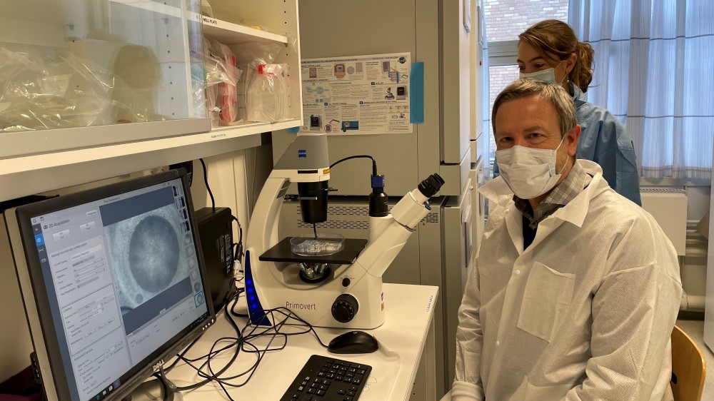 Bildet viser en mann med munnbind foran en dataskjerm med et bilde, og en kvinne med munnbind bak ham ved et mikroskop