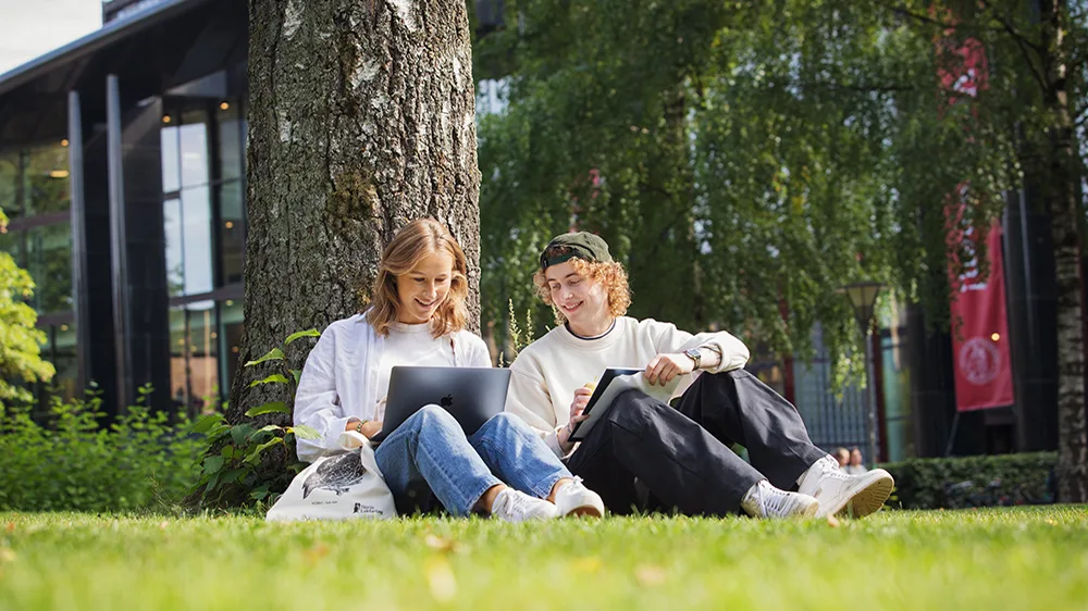 To studenter sitter ute på gressplen på campus, de smiler og ser sammen på en datamaskin