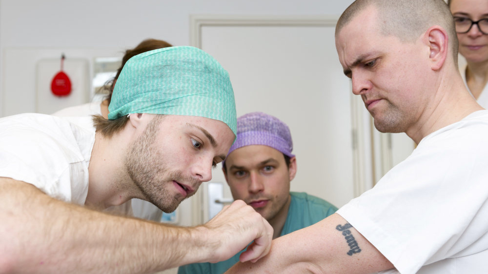 Bilde av en legestudent som undersøker en person som spiller rollen som pasient. To andre personer følger med.