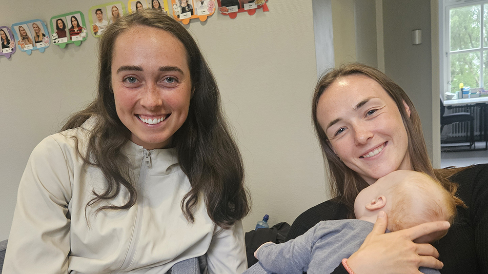 to kvinnelige studenter sitter i en sofa, en av dem holder en baby